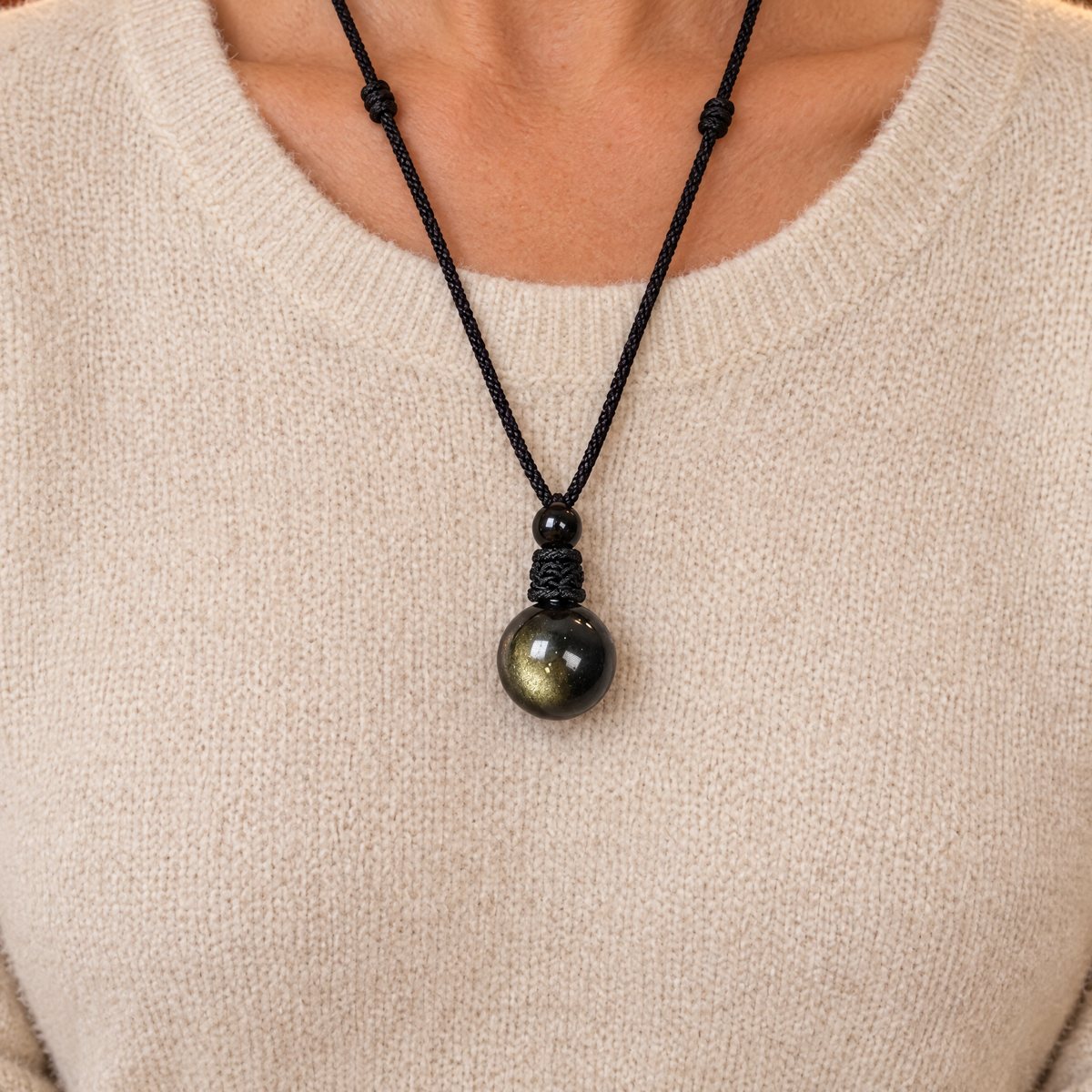 The sphere at the neckline of a woman of about 45 over a cream knit sweater, the cord long, window light, her hand near it